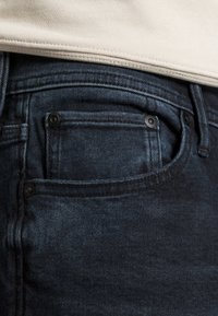 Detailný záber na vrecko tmavomodrých džínsy, s viditeľným šitím a kovovými nitmi, čiastočne zakryté béžovou látkou.