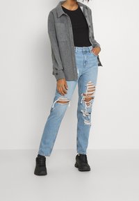 Chemise en flanelle grise portée sur un t-shirt noir, associée à un jean bleu clair déchiré et des baskets noires. Accent mis sur le tissu usé et la coupe décontractée.