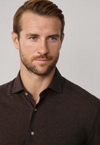 Hombre con barba clara y pelo peinado que lleva una camisa oscura de botones, mirando ligeramente hacia un lado contra un fondo claro y liso.