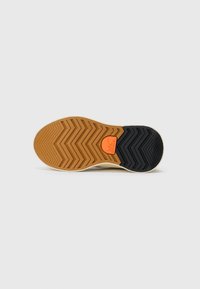 Semelle d'une sneaker montrant un motif en caoutchouc marron et noir avec un petit patch logo orange au milieu sur fond blanc.
