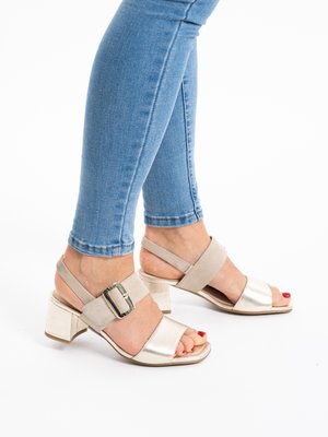 Feet in beige open-toe block heel sandals with buckle straps, paired with light blue skinny jeans, on a white background.
