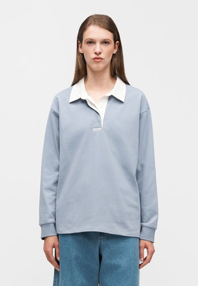 Ανοιχτό μπλε πουλόβερ από μαλακό ύφασμα με λευκό γιακά πόλο, μακριά μανίκια και μικρή ετικέτα με το λογότυπο στο στήθος.
