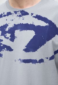 Κοντινό πλάνο ενός ανοιχτογκρι πουκαμίσου με μεγάλο, αφηρημένο σκούρο μπλε σχέδιο από πιτσιλιές χρώματος στην περιοχή του στήθους.