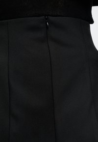 Gros plan sur une jupe noire texturée portée avec un haut noir, montrant une fermeture éclair dissimulée sur le côté de la taille.