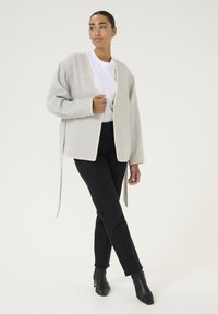 Cardigan grigio chiaro con rifiniture a contrasto, apertura frontale e maniche lunghe. Abbinato a jeans neri e stivaletti alla caviglia. Texture liscia.