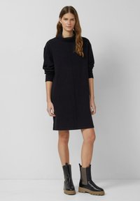 Czarna sukienka typu sweatshirt z kołnierzem typu komin, długimi rękawami i prostym krojem. Noszona z czarnymi botkami za kostkę z teksturowanymi bocznymi panelami.