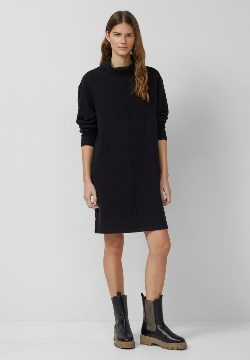 Czarna sukienka typu sweatshirt z kołnierzem typu komin, długimi rękawami i prostym krojem. Noszona z czarnymi botkami za kostkę z teksturowanymi bocznymi panelami.