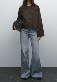 Kvinna iklädd en lös mörkbrun jacka och vida ljusblå jeans, som håller en liten svart handväska mot en enfärgad bakgrund.