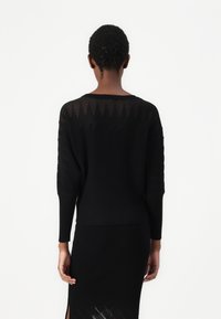 Melns struktūruots adījuma džemperis ar garām piedurknēm, ribotu dizainu un caurspīdīgu aizmuguri. Apvienots ar saskaņotu melnu svārku, kuram ir šķirba.