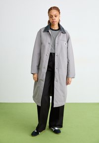 Cappotto lungo grigio con colletto scuro, chiusure a bottone e taschino sul petto. Indossato su una camicia a righe bianco e nere e pantaloni neri.