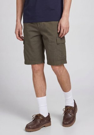 Uomo che indossa pantaloncini cargo verde oliva, calzini bianchi, scarpe di pelle marrone con lacci e una camicia blu navy, in piedi su un pavimento grigio chiaro.