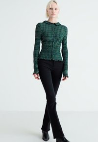 Top de manga larga verde con un patrón, cierre delantero de botones, diseño ajustado y puños con volantes, combinado con pantalones de campana negros y botas de punta negras.