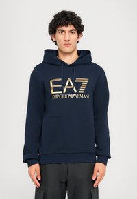 Marineblå hettegenser med gull "EA7 Emporio Armani" logo, frontlomme i kenguru-stil og ribbestrikkede mansjetter og nederkant. Myk tekstur.