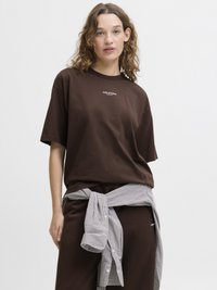 Bruine katoenen T-shirt met korte mouwen, voorzien van een klein wit logo. Een geruite blouse is om de taille geknoopt.