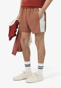 Pantalones cortos marrones con cintura elástica y paneles laterales blancos, con un pequeño logo verde. Combinados con zapatillas crema y calcetines a rayas.