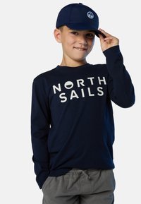 Camisa de manga larga azul marina con el texto blanco "NORTH SAILS"; combinada con una gorra azul marina que presenta un logotipo redondo. El modelo lleva pantalones cortos grises.