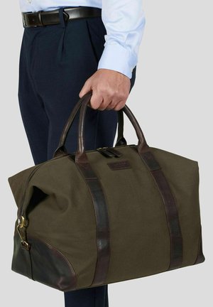 Mann i blå skjorte og mørke bukser holder en stor helgrønn og brun weekend-reiseveske i skinn ved håndtakene.