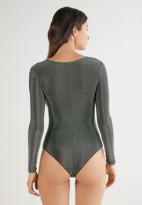 Bodysuit de manga larga en tejido gris metálico. Presenta un escote redondo y una forma ajustada, con una pierna de corte alto y detalles de hardware mínimos.
