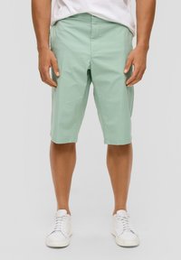 Homme portant un short vert clair jusqu'aux genoux et des baskets blanches, se tenant devant un fond gris clair uni.