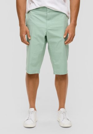 Mann trägt hellgrüne knielange Shorts und weiße Turnschuhe und steht vor einem einfarbigen hellgrauen Hintergrund.