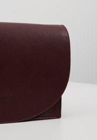 Borsa a tracolla in pelle bordeaux con finitura testurizzata, chiusura a patta arrotondata e linee pulite. Bordo liscio e costruzione solida.