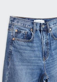 Jean en denim bleu avec bouton devant, passants de ceinture et petite poche avant en gros plan.