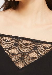 Czarna odzież z trójkątnym wstawkiem koronkowym o skomplikowanych wzorach kwiatowych, uzupełniającym gładki materiał poniżej.