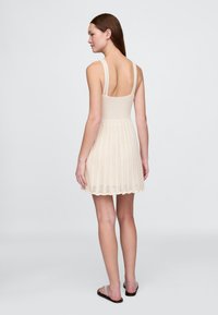 Stickad krämfärgad klänning med figursydd överdel och utsvängd fåll med vågformad kant. Har breda axelband och öppen rygg, texturerat mönster på kjolen.