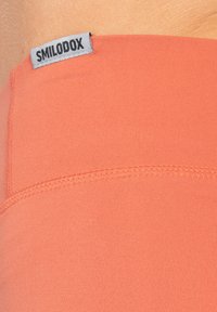 Tessuto sportivo color corallo con superficie liscia. Cuciture in vista e un'etichetta grigia a marchio "SMILODOX" sulla vita.
