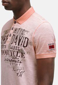 Poloshirt in lichtroze met een textuurstof. Heeft een knoopsluiting en printdetails in donkerbruin, evenals een patch op de mouw.