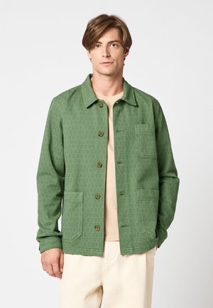 Homme portant une veste verte boutonnée avec trois poches avant par-dessus une chemise beige et un pantalon de couleur claire, sur un fond uni.