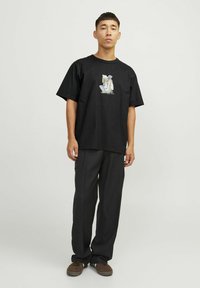 Camiseta de algodón negra con un estampado gráfico en la parte delantera, combinada con pantalones sueltos negros y zapatos de ante marrón.