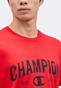 Άνδρας με κόκκινη μπλούζα με τη λέξη "CHAMPION" και το λογότυπο τυπωμένα μαύρα μπροστά.
