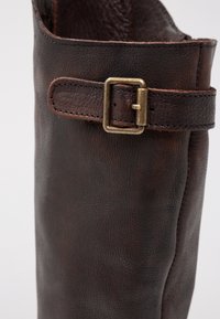 Bota de piel marrón oscuro con un acabado texturizado, construcción robusta y un acento de hebilla de latón en la correa ajustable alrededor de la parte superior.