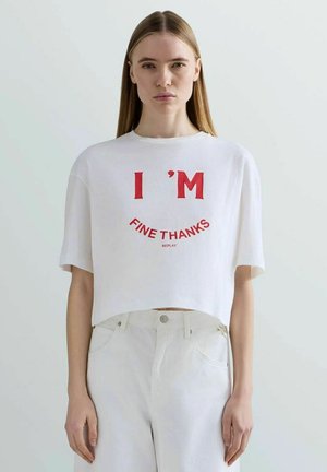 Donna con capelli biondi lisci indossa una maglietta corta bianca con testo rosso "I'M FINE THANKS" e pantaloni bianchi a vita alta.