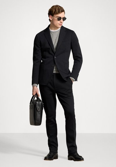 Blazer noir sur mesure, pull gris, pantalon noir et chaussures en cuir. Tenant un sac en cuir noir, avec des lunettes de soleil rondes. Design simple et moderne.