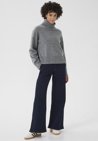 Grauer Rollkragenpullover aus weichem Material, kombiniert mit lockeren, marinen Schlagjeans und weißen Sneakers mit schwarzen Streifen.