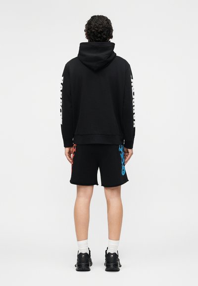 Person iført svart hettegenser med teksten «PARROW» på ermene og svarte shorts med røde og blå flammemønstre, stående med ryggen mot, mot en hvit bakgrunn.