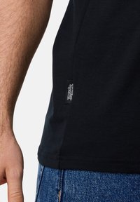 Sort bomulds T-shirt med rund hals, sidesøm og et lille mærkelabel nær kanten, kombineret med blå denim jeans.