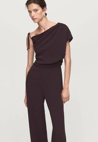 Bordeaux off-shoulder jumpsuit met gedrapeerde stof, wijde pijpen en een vastgebonden schouderdetail. Heeft een gladde textuur en een minimalistisch ontwerp.