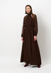 Bruine jurk met lange mouwen van lichte stof, met een hoge halslijn en een geplooide taille, en transparante mouwen en een zijsplit.