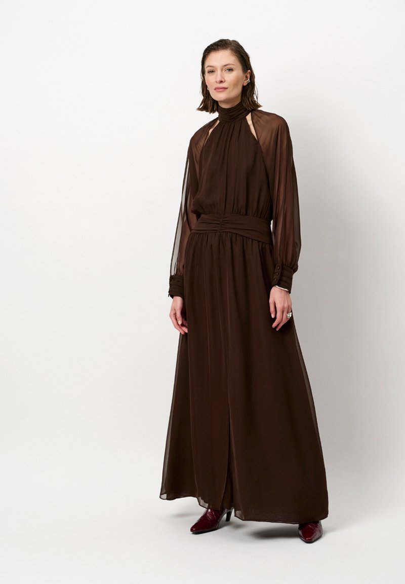 Bruine jurk met lange mouwen van lichte stof, met een hoge halslijn en een geplooide taille, en transparante mouwen en een zijsplit.
