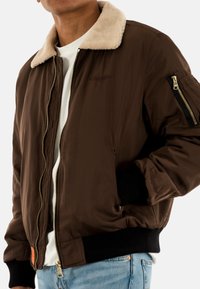 Braune Bomberjacke mit einem Kragen aus Kunstpelz, Reißverschlussfront, Seitentaschen und gerippten Bündchen. Verfügt über ein strapazierfähiges Außenmaterial und goldene Akzente.