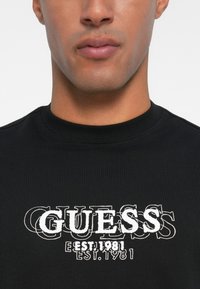 Sudadera negra con cuello acanalado, que presenta el texto "GUESS" en blanco con un efecto en capas y "EST. 1981" debajo.