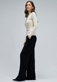 Blusa de manga larga color crema con detalle drapeado, combinada con pantalones negros de pierna ancha de talle alto y tacones negros puntiagudos sobre un fondo neutro.