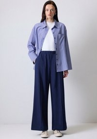 Camisa de botones a rayas azul y blanca, camiseta blanca, pantalones con cinturilla elástica en navy y zapatillas beige. Modelo de pie con las manos en los bolsillos.