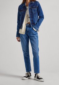 Denimjacka i mörkblått med framfickor, kombinerad med ljusblå jeans och svarta sneakers med vita detaljer. En bomullströja synlig underneath.