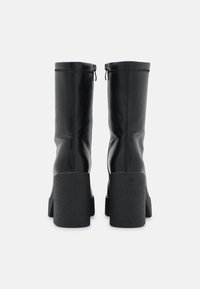 Bottes noires en cuir montantes jusqu'au genou avec un talon noir texturé et épais. Elles comportent une fermeture éclair et une couture verticale distinctive sur la tige.