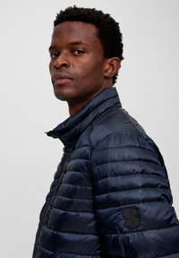 Man met kort krullend haar die een marineblauwe gewatteerde jas met een hoge gegroeide kraag draagt, terwijl hij over zijn schouder kijkt tegen een neutrale achtergrond.