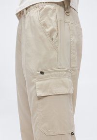 Μπεζ παντελόνι cargo με τσέπες με φερμουάρ και καπάκια, ελαστική μέση και πλαϊνές θηλιές, απεικονίζεται από το ισχίο μέχρι το μέσο του μηρού σε φωτεινό φόντο.